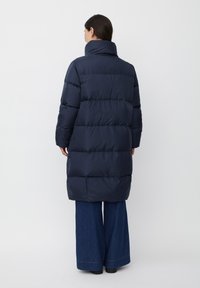 Giacca imbottita blu navy con colletto alto, design trapuntato e maniche aderenti, che arriva a metà coscia. Indossata sopra pantaloni in denim svasati.