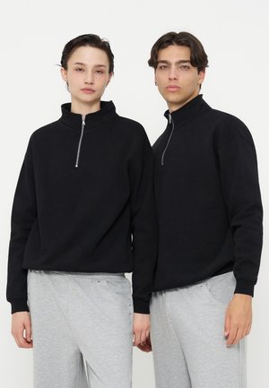 Deux personnes portant des sweat-shirts noirs à fermeture éclair quart et des pantalons gris clair se tiennent côte à côte devant un fond blanc uni.