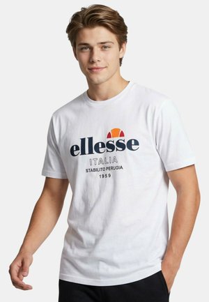 Jeune homme portant un t-shirt blanc ellesse Italia avec logo et texte, debout avec une main dans la poche devant un fond uni.