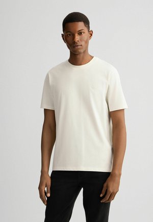 Hvid bomulds T-shirt med rund hals og korte ærmer, med et diskret logo på brystet. Båret med sorte jeans.
