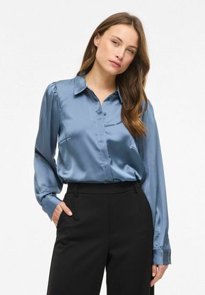 Kobieta o długich brązowych włosach, ubrana w niebieską satynową bluzkę z długim rękawem, wpuszczoną w czarne spodnie z wysokim stanem, stojąca z jedną ręką w kieszeni.