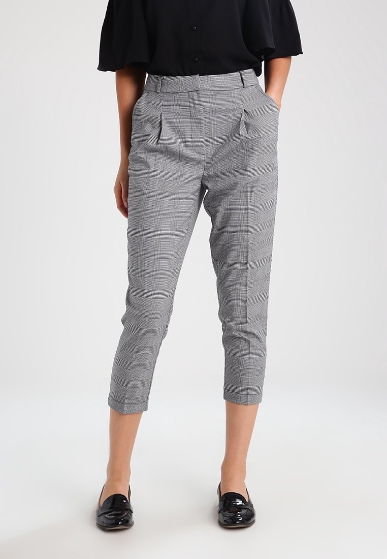 Femme portant un pantalon gris à carreaux, fuselé, longueur cheville, avec des plis à l'avant, associé à des mocassins noirs et un haut noir ample.