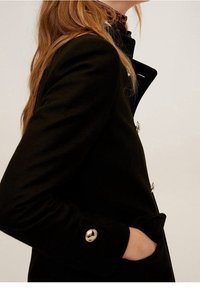 Abrigo negro con un diseño estructurado, que presenta botones plateados brillantes, un cuello alto y una mano en el bolsillo lateral. Textura suave.