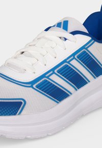 Chaussure de sport blanche avec des accents rayés bleus, lacets blancs, dessus en mesh et semelle rembourrée.