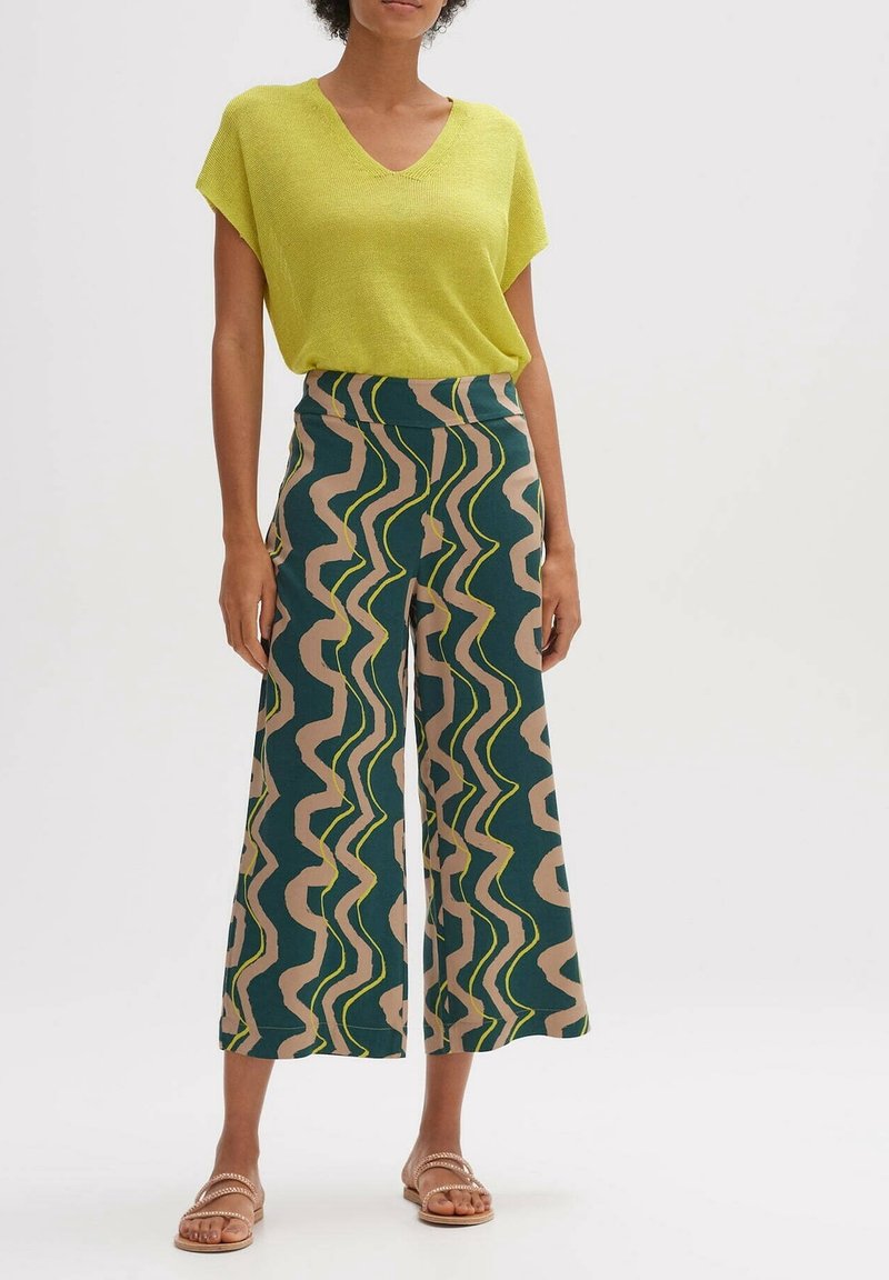 Femme portant un haut manche courte vert citron à col en V et un pantalon ample coupé court avec un motif ondulé vert, beige et jaune, sandales beige.