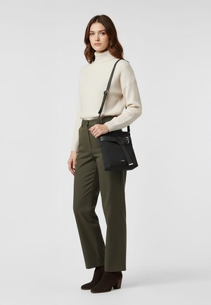Mujer con suéter de cuello alto color crema, pantalones oliva, botines negros y un bolso cruzado negro con detalle de hebilla.