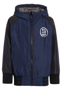 Granatowa kurtka przeciwwiatrowa z szarym podszewką, z zapięciem na zamek, kapturem i białym logo na klatce piersiowej. Czarny akcent na ramionach i mankietach.