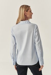 Camicia a maniche lunghe azzurro chiaro con colletto, polsini abbottonati, texture liscia e orlo curvo, vista da dietro.