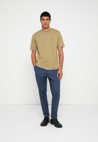 Light olive green short-sleeve t-shirt with a round neckline and a small logo, paired with dark blue trousers and black shoes.
