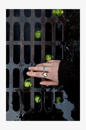 Silver rings on a hand positioned above a dark metal grate with green brussels sprouts scattered in puddles of water.