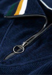 Marinblå zip-jacka med en texturerad yta, som har en silverfärgad dragkedja och flerfärgade randiga detaljer i grön, gul och vit på kragen.