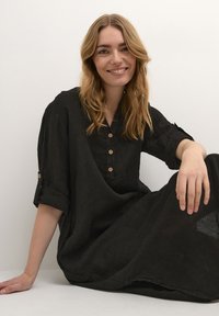 Abito camicia in lino nero con colletto, tre bottoni in legno e maniche arrotolate. Il tessuto ha un aspetto morbido e texture.