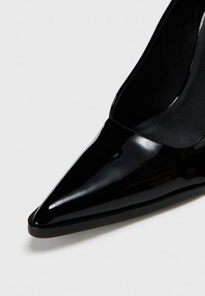 Pantofii cu toc înalt din piele lacuită neagră au vârful ascuțit și o siluetă curbată, având un finisaj neted și lucios, precum și un design minimalist.