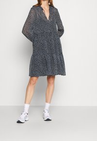 Mujer vistiendo un vestido negro de flores hasta la rodilla con mangas largas y zapatillas blancas, de pie frente a un fondo claro y liso.