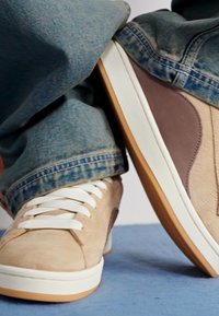 Beige suede sneakers with white laces, brown accents, and rubber soles. Worn with blue denim jeans featuring a slight fade.