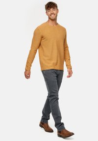 Mustard yellow textured knit sweater paired with gray jeans and brown ankle boots; model walking with a neutral background.
