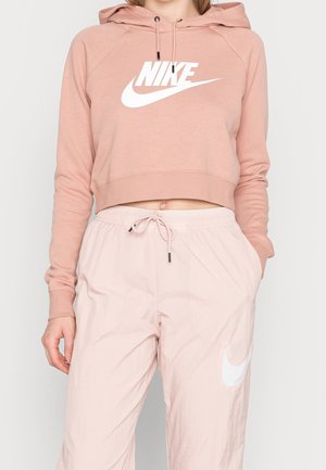 Kobieta nosząca jasnoróżową krótką bluzę Nike z logo oraz pasujące jasnoróżowe spodnie z troczkiem i białym logo Nike na udzie.