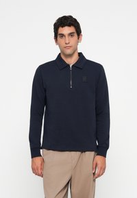 Man wearing navy long-sleeve polo with front zipper and beige pants, standing against a plain white background.