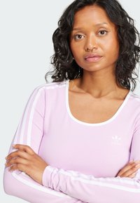 Ljuslila långärmad topp med vita accentfärger, rund halsringning och tre vita ränder som löper ner längs varje arm. Adidas-logotyp i vitt.