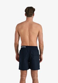 Marineblauwe zwembroek met elastische tailleband en witte "SPORTS" tekst, voorzien van een achterzak met knoopdetail. Glad stof textuur.