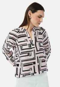 Bluzka z długim rękawem, wykonana z lekkiego materiału, z geometrycznym wzorem w czerni i różu, z częściowo odkrytym dekoltem i luźnym krojem.