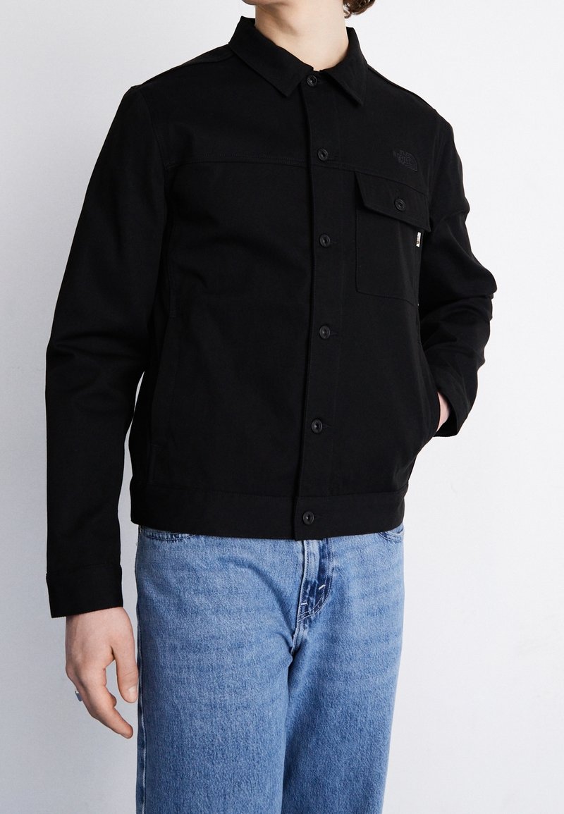 Veste en toile noire avec fermeture à boutons, poche poitrine et détail logo. Associée à un jean en denim bleu clair, affichant une silhouette décontractée.