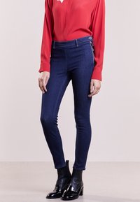 Tmavě modré džíny skinny s vysokým pasem, se bočními zipy, spárované s červenou blůzou s dlouhým rukávem a černými kotníkovými botami.