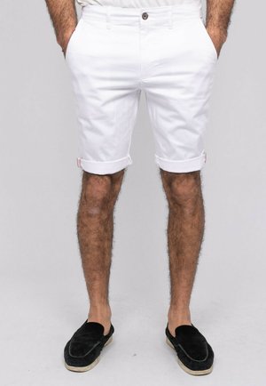 Homme portant un short blanc retroussé avec des poches avant, des chaussures noires sans lacets, les mains dans les poches, sur un fond uni.
