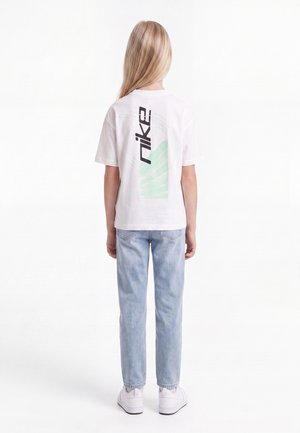 Kind mit langen blonden Haaren, das ein weißes Nike-T-Shirt, hellblaue Jeans und weiße Turnschuhe trägt, steht mit dem Rücken vor einem weißen Hintergrund.