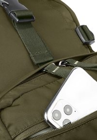 Smartphone avec triple caméra partiellement dans la poche zippée d'un sac à dos en tissu vert olive.