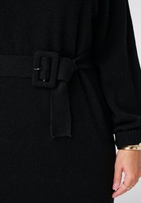 Zwarte gebreide jurk van een gestructureerde stof, met een brede tailleband en lange mouwen, die een ontspannen silhouet creëert.