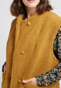 Gilet en polaire jaune moutarde, duveteux, avec un col arrondi, des boutons sur le devant et des poches latérales, superposé à une chemise à motif floral.