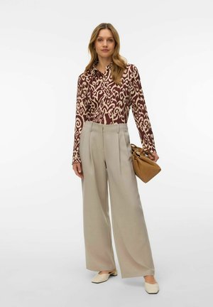 Vrouw gekleed in een bruin-beige blouse met lange mouwen en patroon, beige wijde broekspijpen, crème platte schoenen, en ze houdt een tan handtas vast.