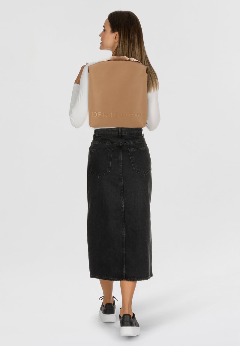 Sac à dos en cuir tan avec logo en relief, de forme rectangulaire et surface texturée. Porté sur le dos d'une personne portant une jupe en denim noire.