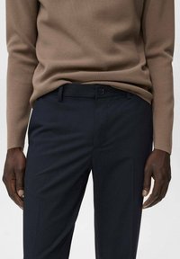 Pull en tricot texturé brun associé à un pantalon de costume bleu marine avec une coupe plate, des passants de ceinture et une fermeture à un bouton.