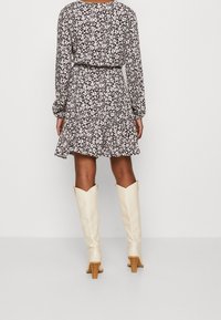 Blumenkleid mit langen Ärmeln, gerafftem Bund, weit ausgestelltem Rock und beigen knielangen Stiefeln mit kleinem Blockabsatz, vor einem neutralen Hintergrund.