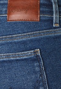 Tela de mezclilla azul con costuras visibles, con una etiqueta de cintura de cuero marrón embosada con "Wrangler". Incluye un diseño estándar de bolsillo trasero.