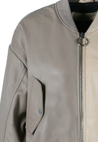 Veste bomber en cuir taupe clair, avec une fermeture éclair argentée, un accent de matériel rond et une poche avec un bouton-pression. Texture lisse.