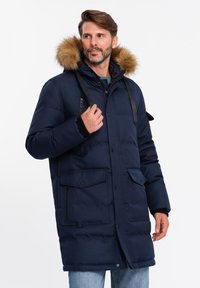 Giacca trapuntata blu navy con cappuccio foderato in pelliccia sintetica, chiusura con cerniera e bottoni a pressione, diverse tasche sul petto e ai lati, e cinghie regolabili.