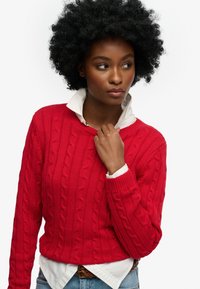 Vrouw met krullend haar, gekleed in een rode kabeltrui over een wit shirt en een blauwe spijkerbroek, kijkt naar rechts met haar hand bij de kraag.