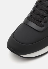 Černé tenisky s hladkým syntetickým svrškem, zaoblenou špičkou, silnou bílou mezipodešví a plochými černými tkaničkami. Texturované detaily na jazyku.