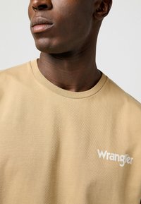 Camiseta de algodón beige con cuello redondo, que presenta un logo blanco de "Wrangler" en el pecho. Textura suave con elementos de diseño mínimos.