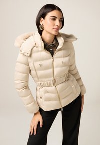 Beżowa kurtka puchowa z kapturem wyłożonym futrem, zapinana na zamek, z paskiem w talii i pikowaną fakturą. Połączona z kwiecistą koszulą z kołnierzykiem oraz czarnymi spodniami.