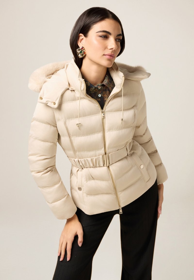 Beżowa kurtka puchowa z kapturem wyłożonym futrem, zapinana na zamek, z paskiem w talii i pikowaną fakturą. Połączona z kwiecistą koszulą z kołnierzykiem oraz czarnymi spodniami.