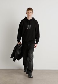 Felpa nera con tasca frontale e dettagli grafici, abbinata a jeans neri e sneakers nere. Modello che indossa una giacca nera. Sfondo semplice.
