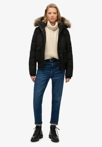 Sort pufferjakke med pelsforet hætte, beige strikket turtleneck, blå lige ben jeans og sorte snørebånd ankelstøvler.