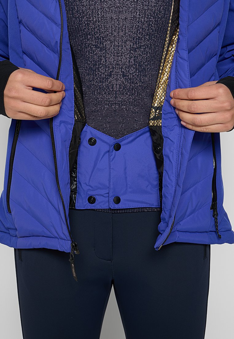 Veste isolée bleue avec un design matelassé, fermeture éclair à l'avant et boutons-pression. La couche intérieure présente un tissu tricoté métallisé texturé.