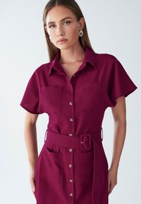 Fuksia nupuga riietus, millel on krae, lühikesed varrukad, ees taskud ja vöökoht. Sile kangas ühtlase värvi viimistlusega.