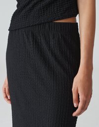 Schwarzer, texturierter Rock mit einem gerafften Taillenbund und einem gekräuselten Stoffdesign, seitlich präsentiert mit einer Hand, die in der Nähe der Naht ruht.