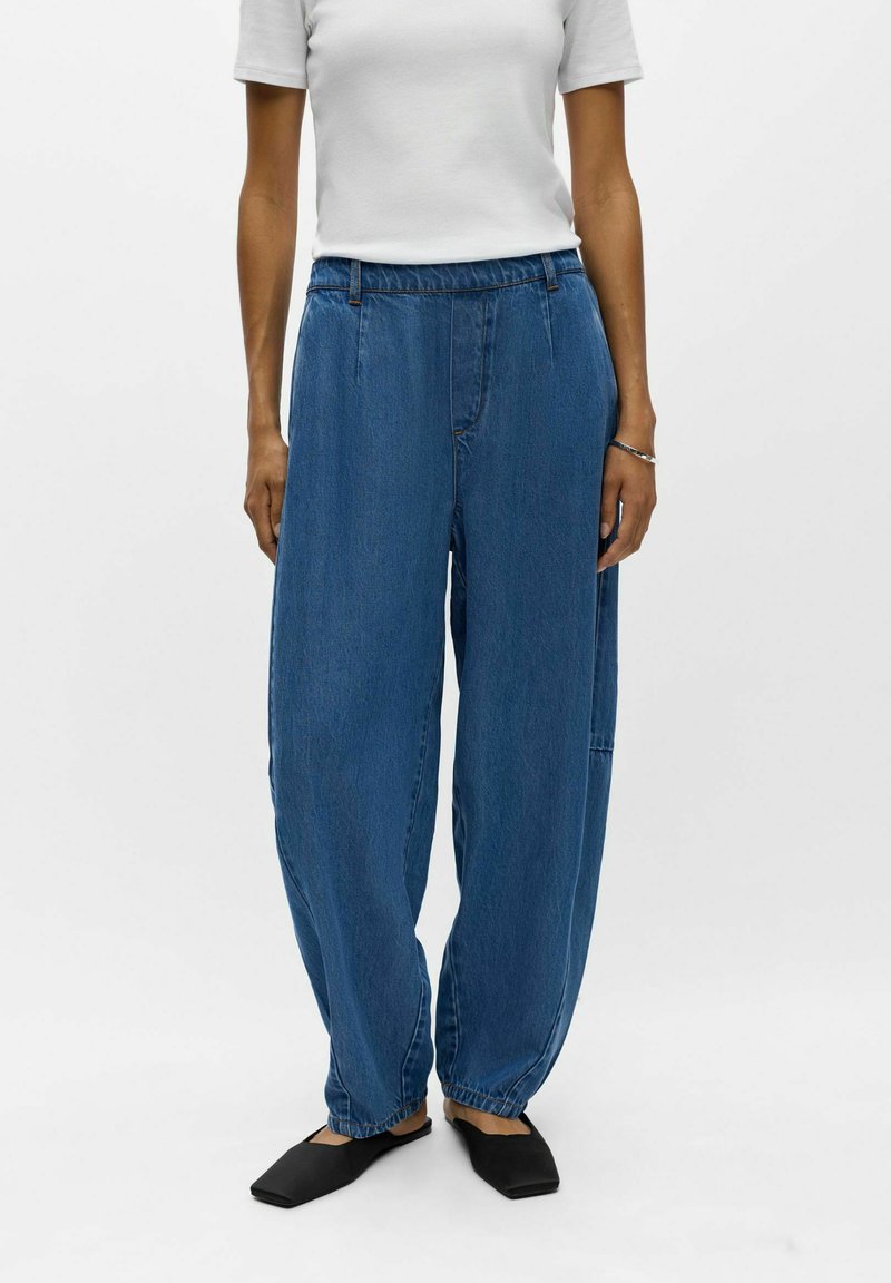 Personne portant un pantalon en jean bleu large avec des poignets élastiques, une chemise blanche à manches courtes, des ballerines noires pointues et un bracelet argenté au poignet droit.